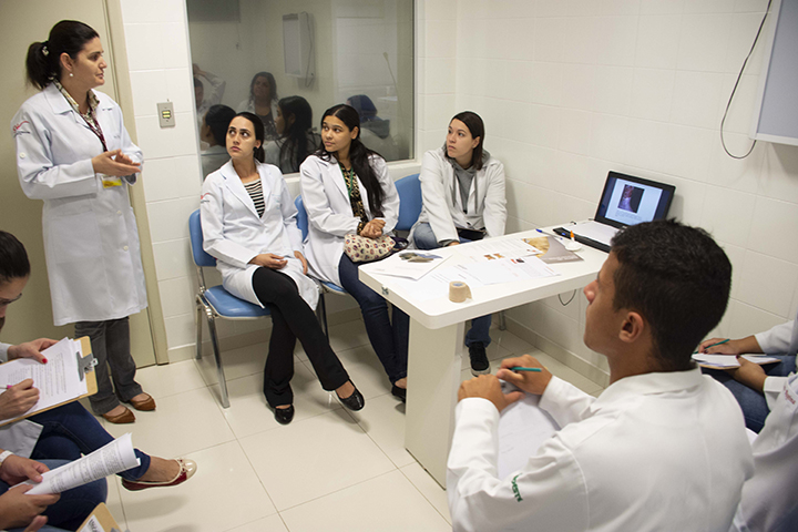 Faculdade Enfermagem: uma pessoa em pé e outras sentadas em um laboratório