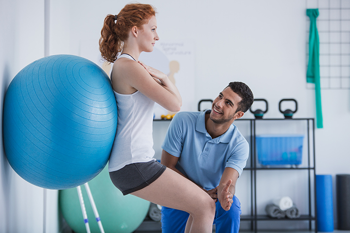 O que faz o fisioterapeuta: Mulher faz exercício de agachamento apoiada em uma bola de pilates na parede e auxílio de um fisioterapeuta