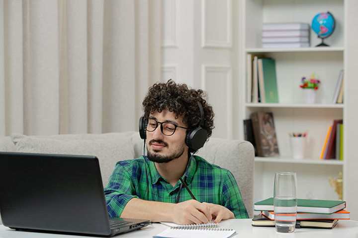 Na foto, um estudante on-line bonito jovem estudando no computador por meio de sites sobre engenharia. Ele usa óculos de grau, fones de ouvido e camisa xadrez verde. O cabelo dele é preto e encaracolado, e ele ele tem a pele branca.